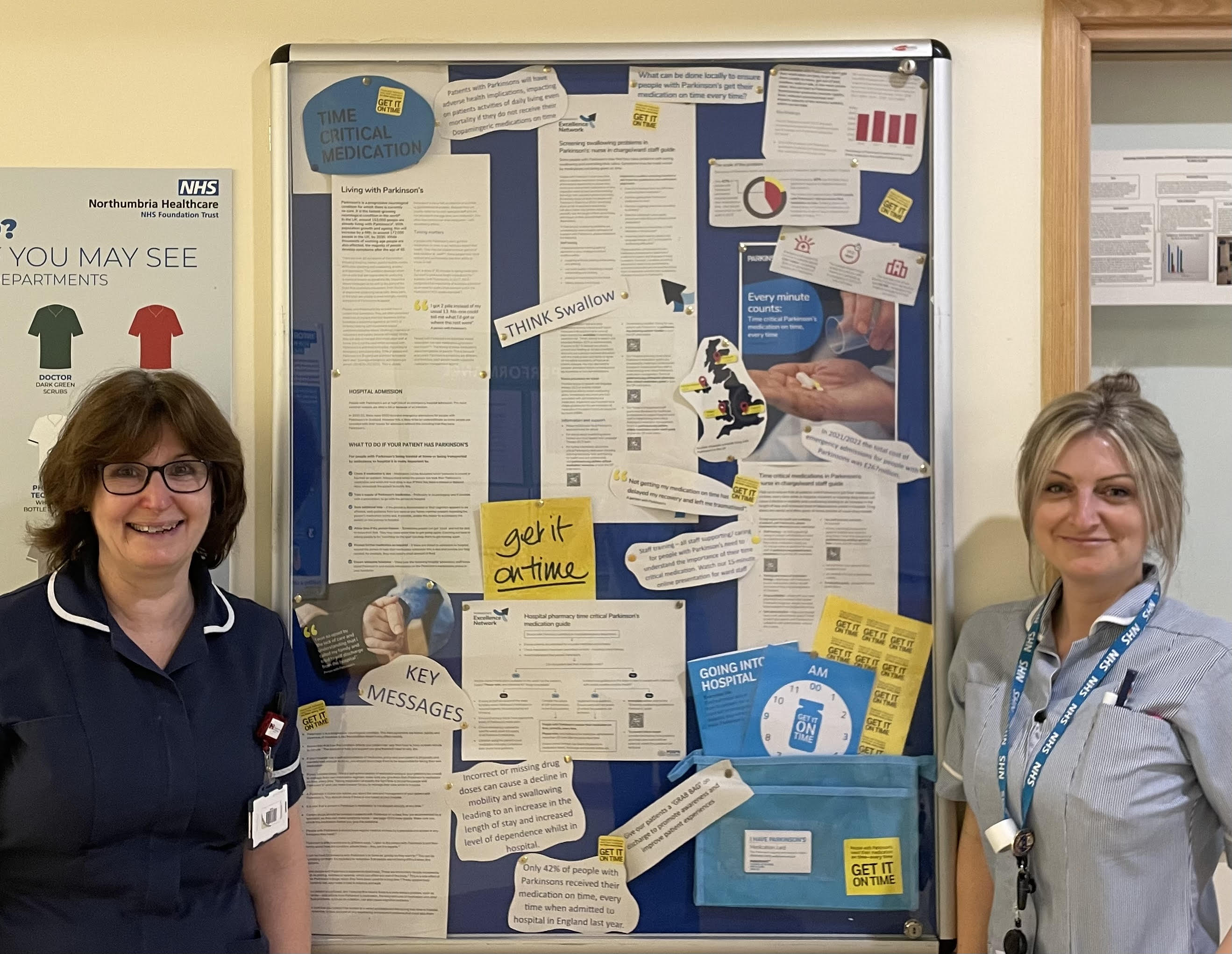 Two nurses stand on either side of a poster display