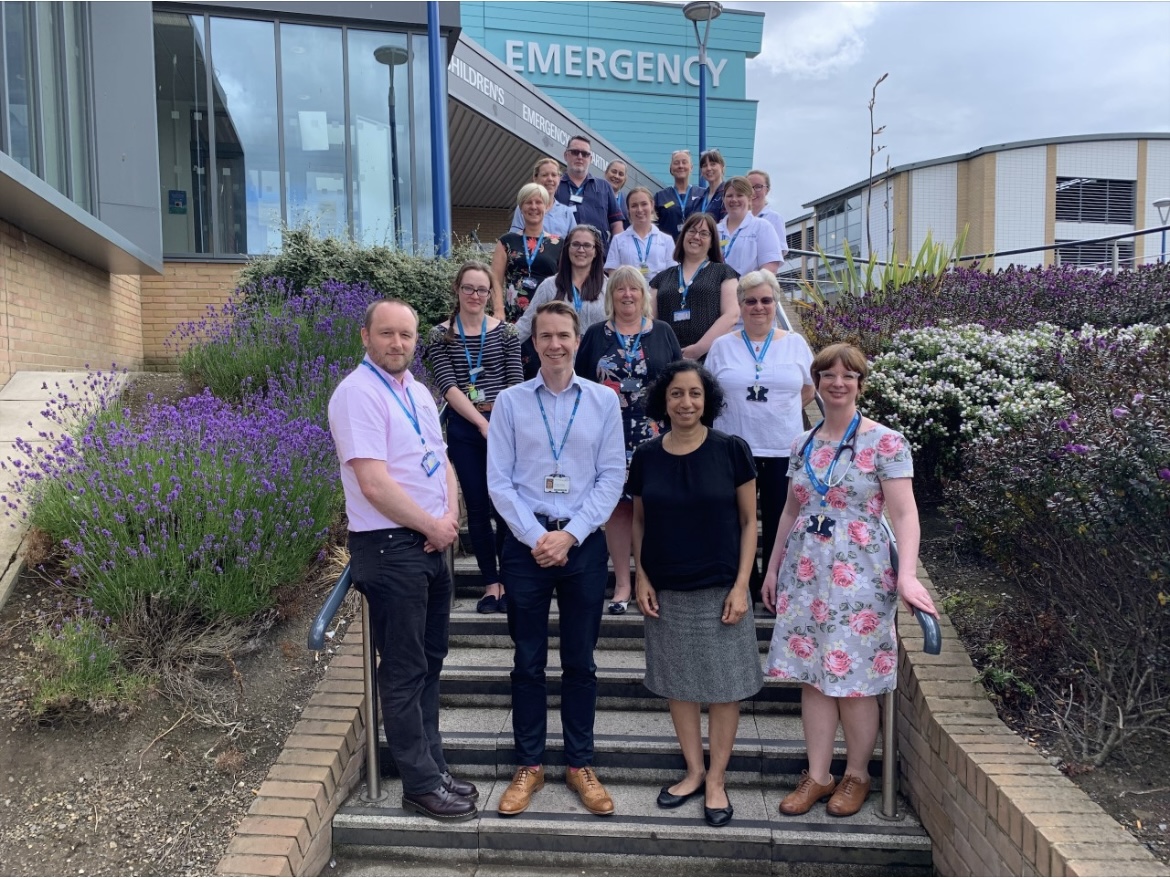 A group of NHS employees stand on steps in front of a hospital emergency department.