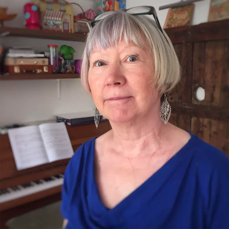 A lady smiles gently at the camera in her house by her piano