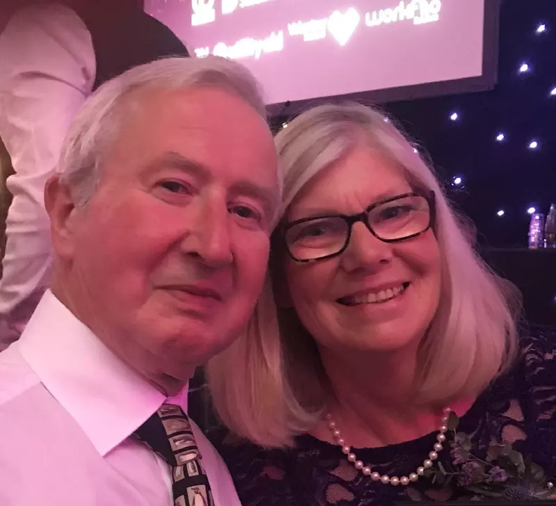 Jim and Jane at an awards ceremony. They are both smiling at the camera. Jim has short white hair and is wearing a white shirt with a black and white patterned tie. Jane has shoulder-length grey hair and is wearing black-rimmed glasses. She is wearing a navy lace dress with a string of pearls. 