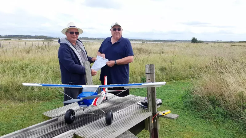 Peter receives his certificate from the assessor. They are standing in a field with a model aircraft in front of them.