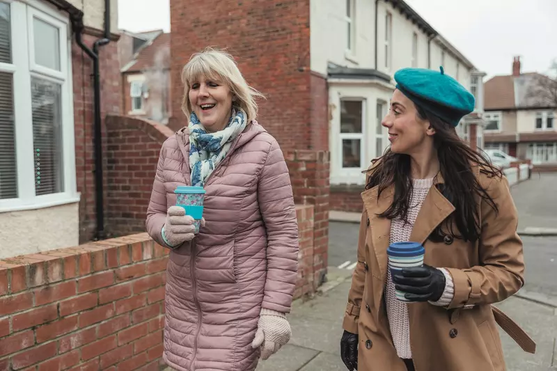 Two ladies walking along the road in winter coats. Both are holding coffee cups.