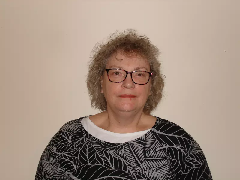 Shirley is smiling at the camera. She has shoulder length hair, and wears glasses. She is wearing a black and white top.