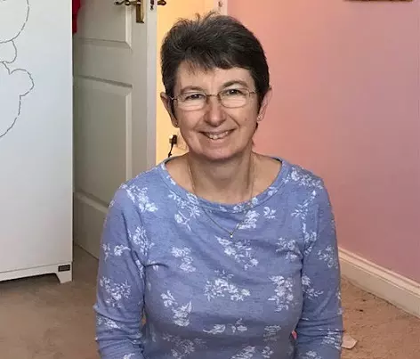 Jayne has short hair and wears glasses. She wears a lilac top and is smiling to the camera.