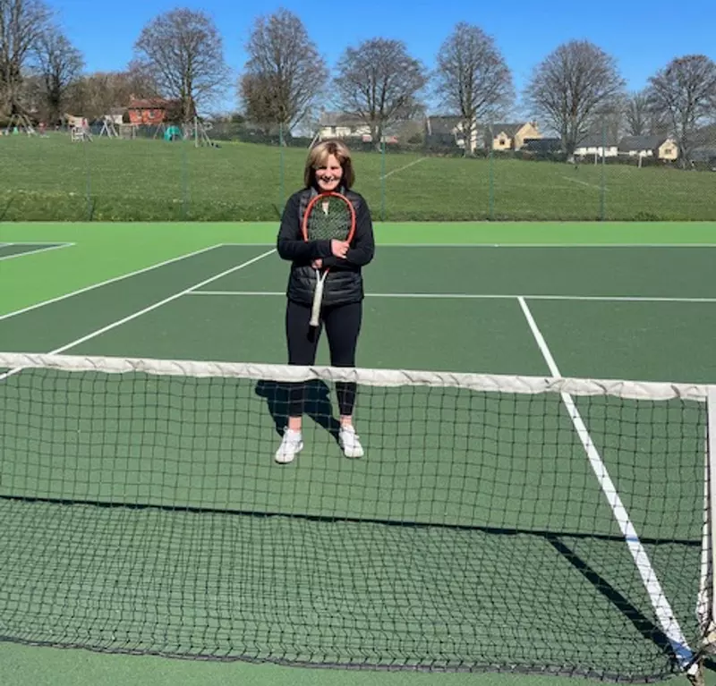 Sharon is stood on a tennis court in front of the net. She is wearing black sports gear and holding a tennis racquet. 