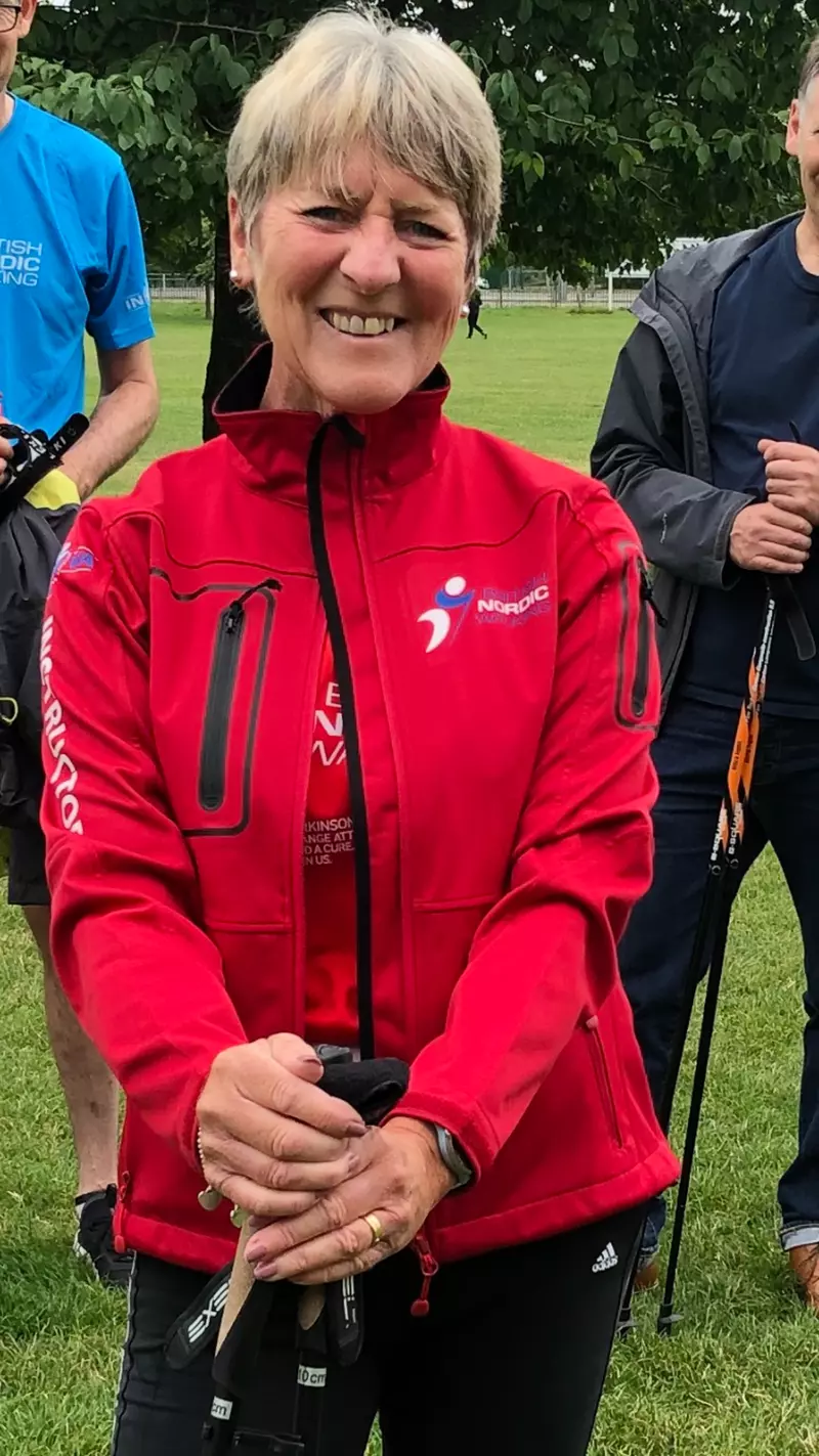An image of a female Nordic walker holding Nordic walking poles and wearing a red walking jacket and black tracksuit trousers