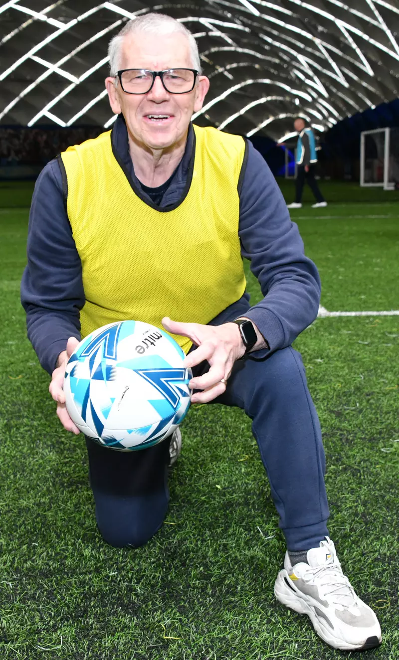 An image of Dave Logan, a walking footballer with Parkinson's wearing a blue t-shirt and blue tracksuit trousers, white trainers and a yellow bib. He is kneeling down in the image while holding a football